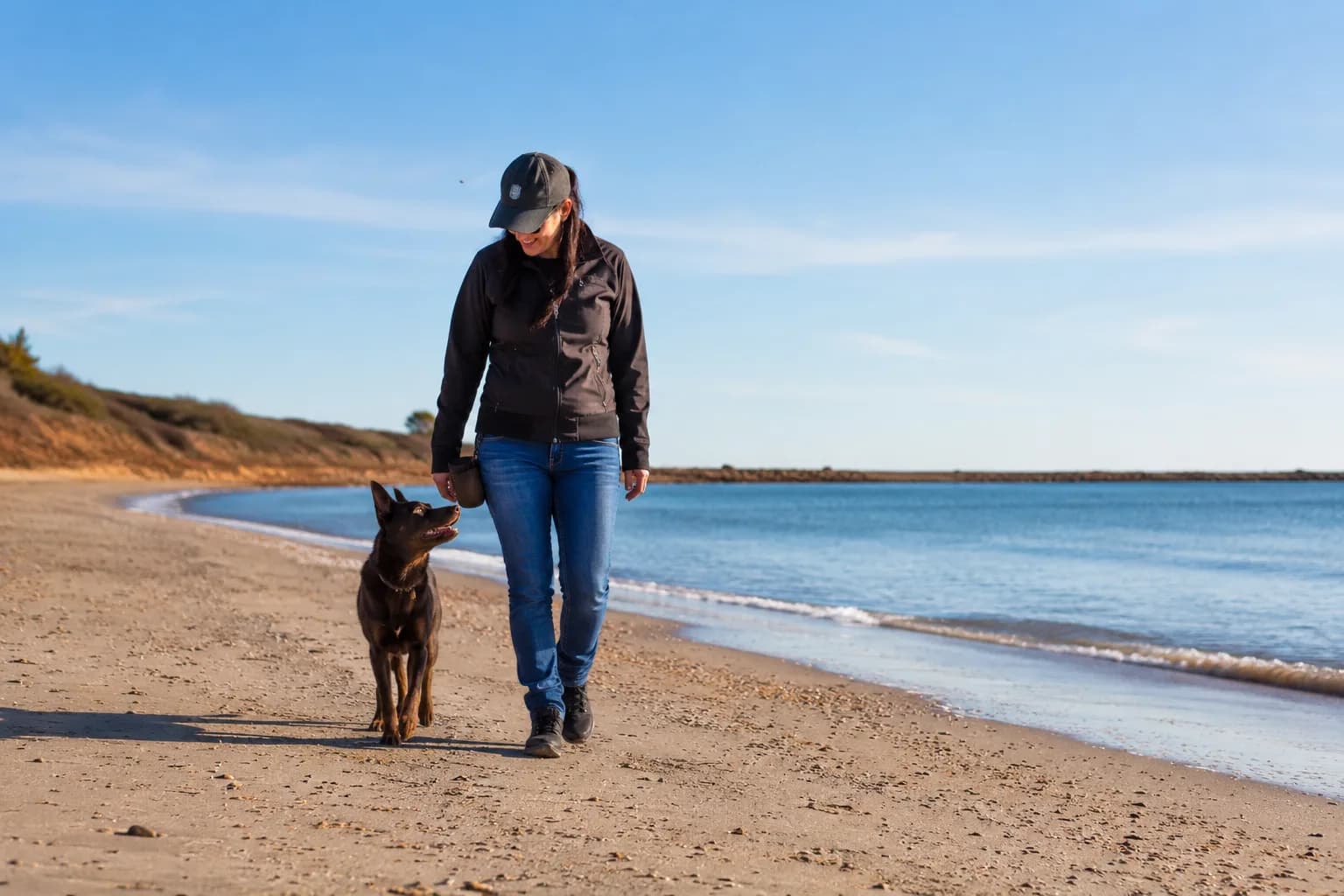 Pauline walking off-lead along a calm regional SA beach with a red kelpie tracking at her side — the picture of reliable recall and connection