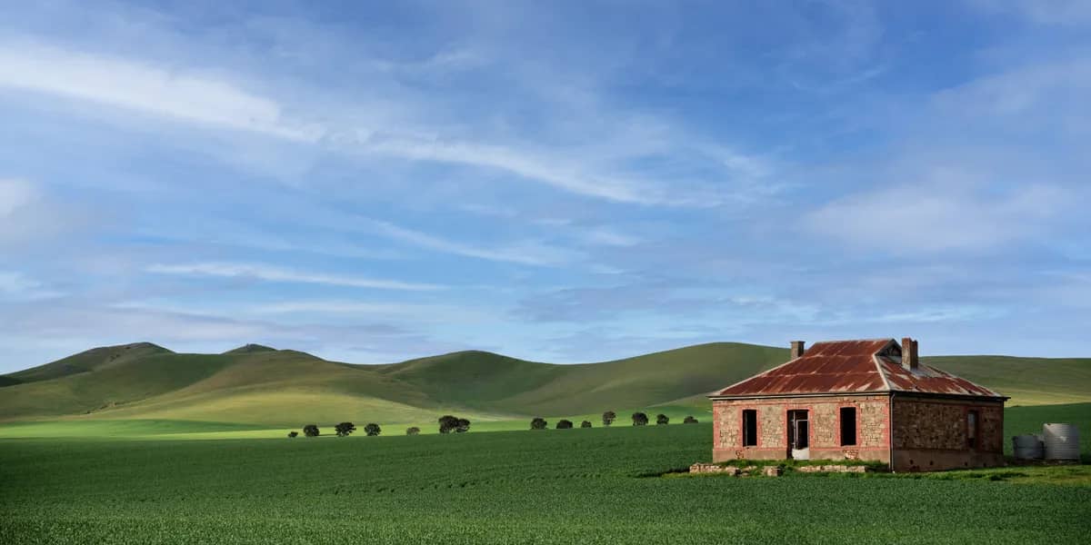 Green wheat paddock with an old stone farmhouse and red-iron roof against the rolling Mid North hills